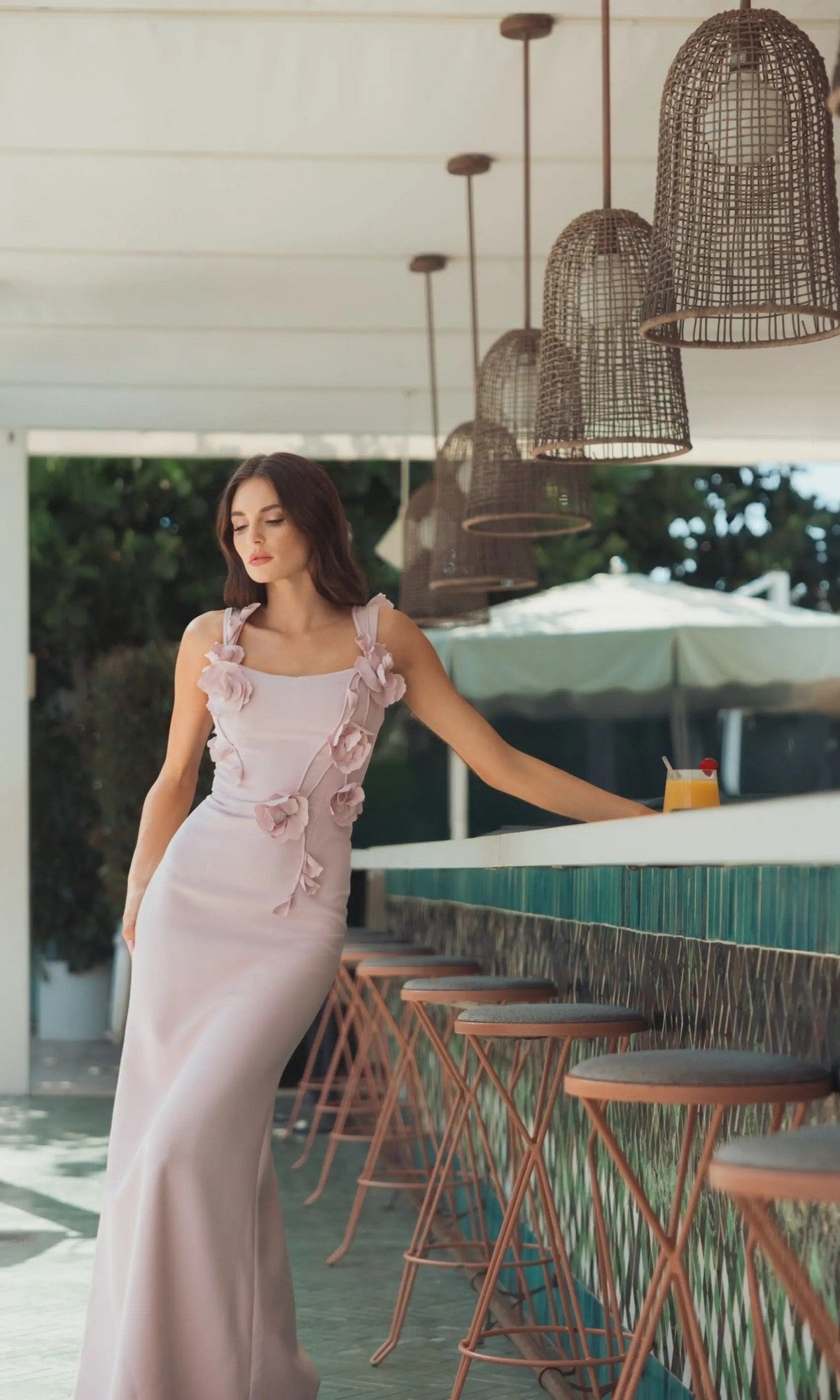 A woman in the Long Formal Dress: Mac Duggal 21236, a light pink gown with floral embellishments, stands by a bar with green tiles, next to a drink under wicker pendant lights.